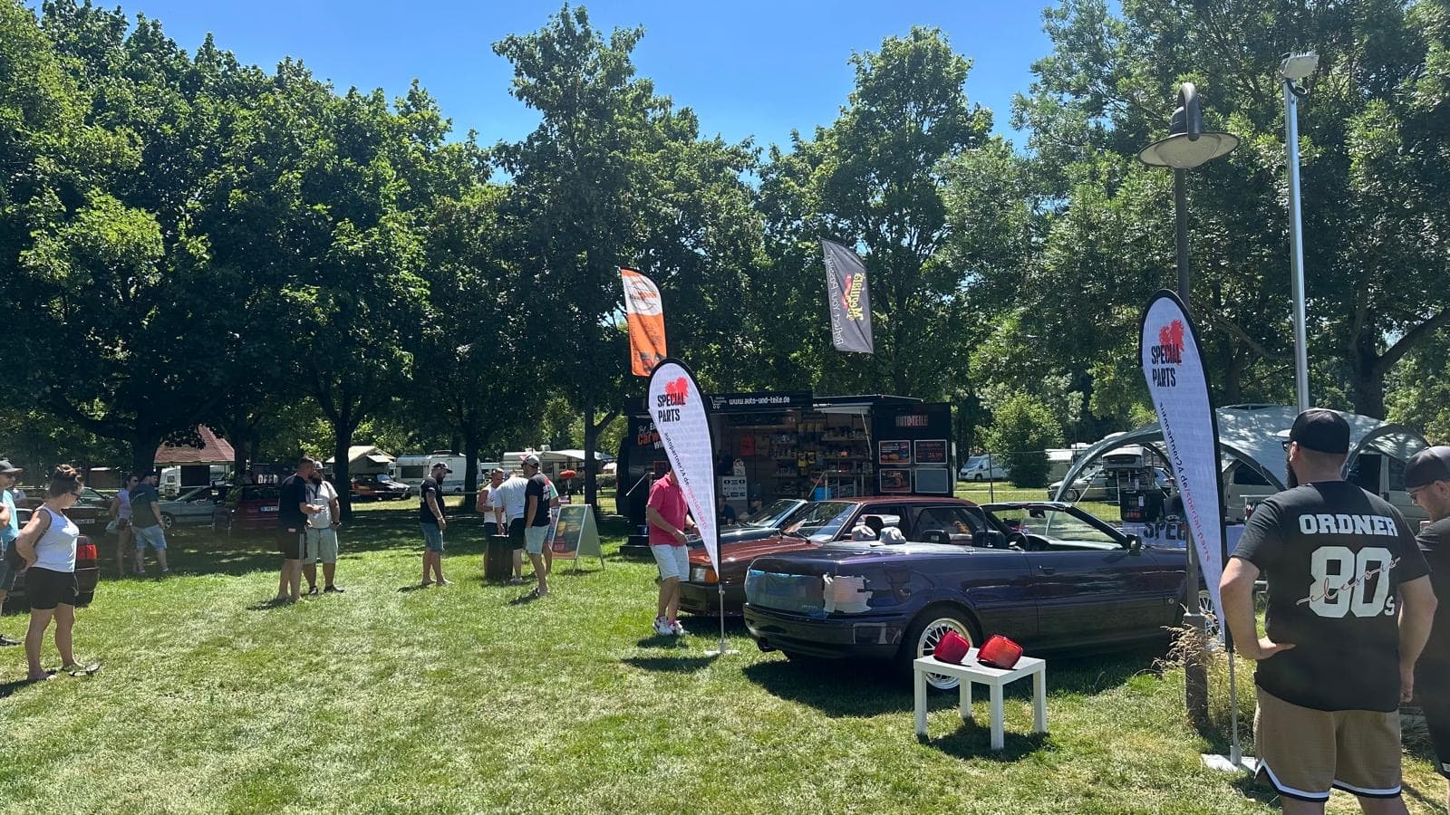 Outdoor car show with a convertible on display, surrounded by people and banners. Trees and a food truck are in the background.