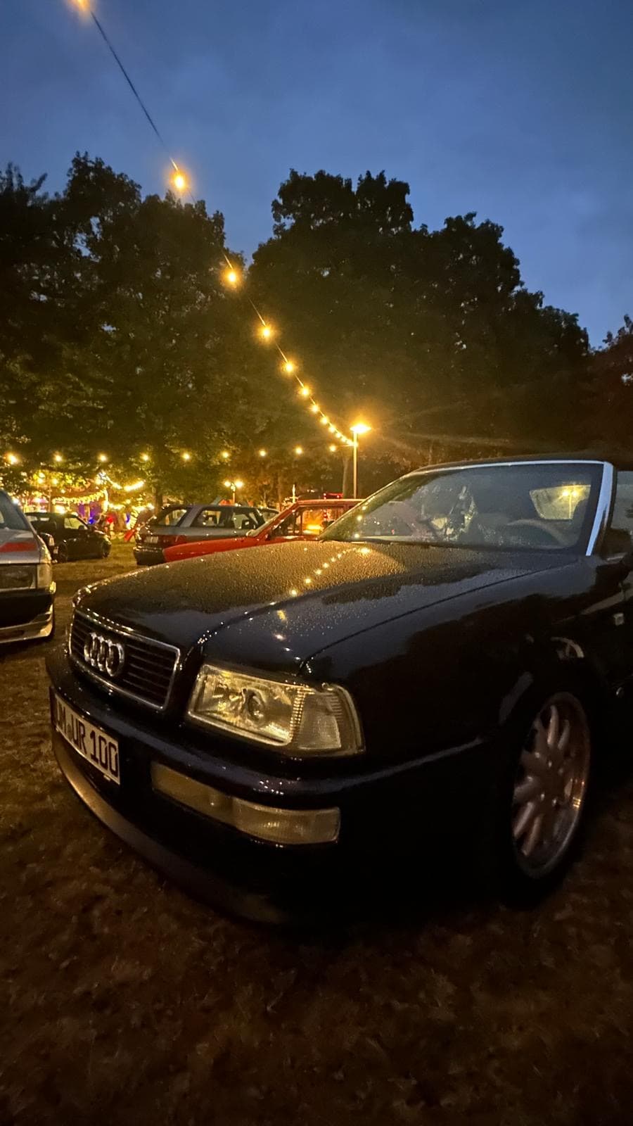 Classic black Audi convertible parked outdoors at dusk, with string lights overhead and trees in the background.