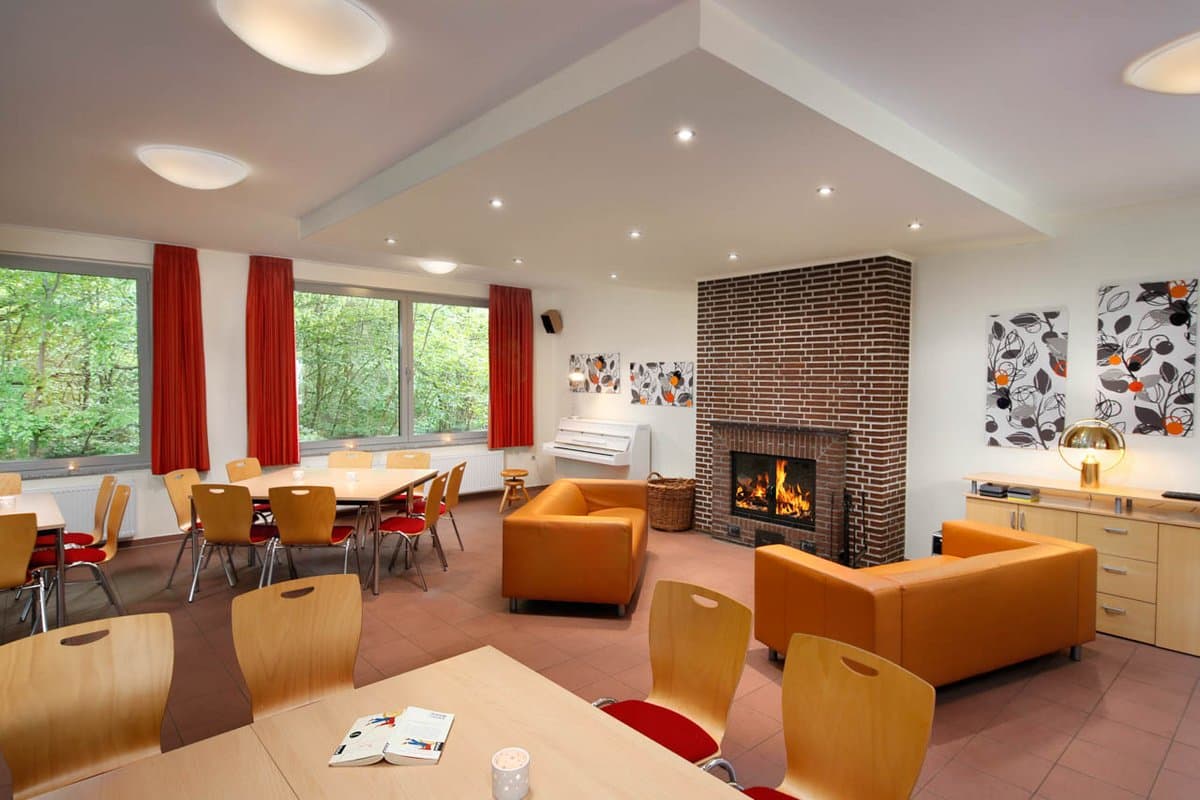 Aufenthaltsraum Gemütlicher Raum mit orangefarbenen Sofas, einem gemauerten Kamin, Tischen, Stühlen und großen Fenstern mit roten Vorhängen mit Blick ins Grüne.