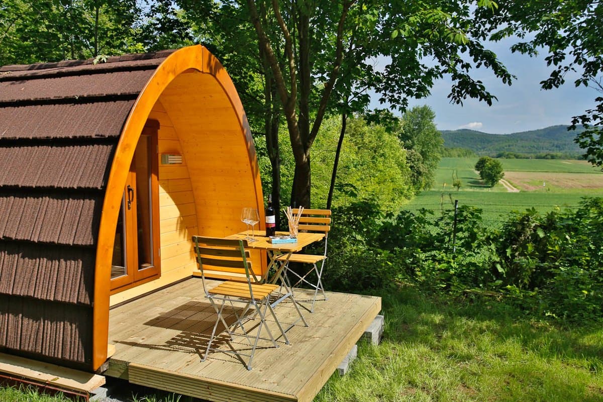 CampingPOD außen Eine gemütliche Holzhütte mit einer kleinen Terrasse, Tisch und Stühlen bietet einen Blick auf eine üppige grüne Landschaft und ferne Hügel unter einem klaren Himmel.