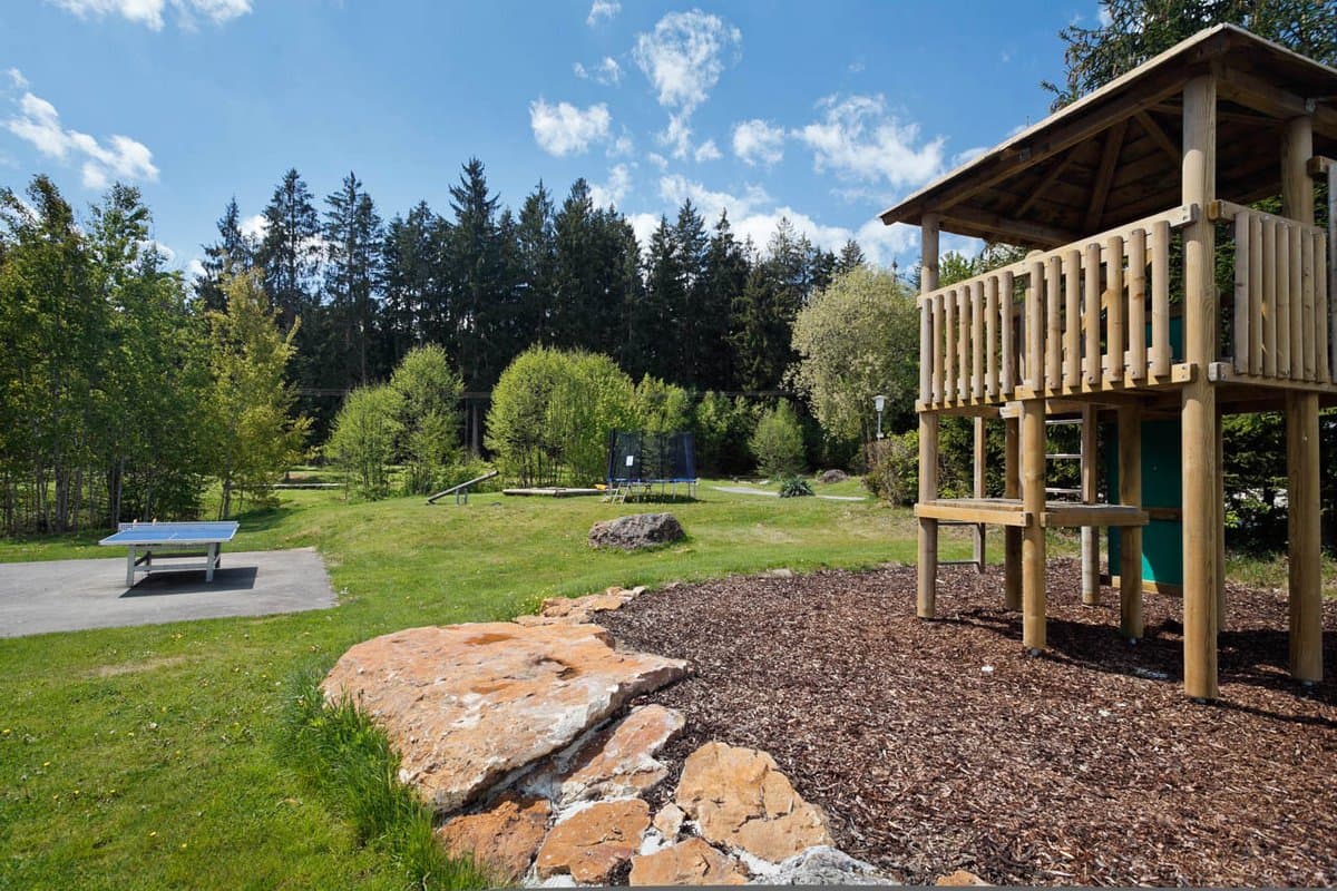 Spielplatz Spielplatz mit einem Holzspielgerät, umgeben von Gras und Bäumen, unter einem blauen Himmel mit Wolken. In der Nähe befindet sich eine Tischtennisplatte.